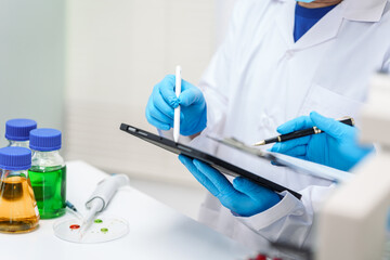 Two Asian scientists in lab coats sit at a table in a laboratory, examining samples through a microscope. The room is equipped with scientific tools, liquids, and chemistry materials.