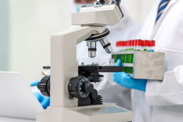 Two Asian scientists in lab coats sit at a table in a laboratory, examining samples through a microscope. The room is equipped with scientific tools, liquids, and chemistry materials.