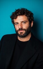Fototapeta premium A handsome man with a beard and curly hair poses against a blue backdrop. He wears a black shirt and jacket.