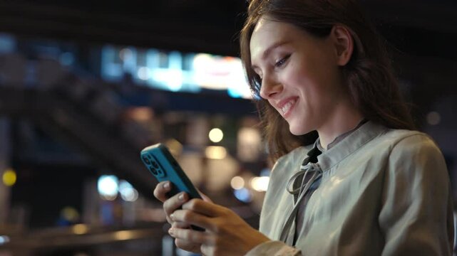 Beautiful Caucasian Smiling Girl Walks Down the Night Street and Texts, Smiling at Her Phone.