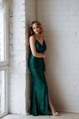 A woman in a green sequined dress is standing in front of a white wall. She is wearing a belt and has her hands on her hips. The dress is long and has a split in the middle