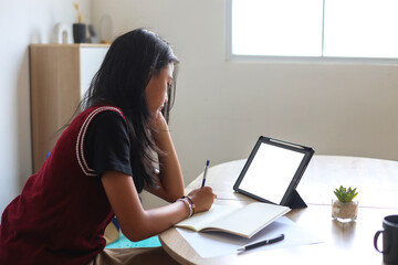 Young Student Girl Wearing School Uniform Studying At Home Using Digital Tablet While Writing On Book
