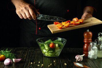 The cook prepares a vegetable mix for dinner. The chef hands with a kitchen board and a knife add sliced cherry tomatoes to the vegetable salad