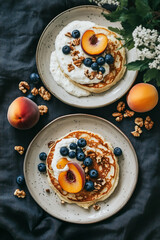 Top view of two plates with cheese pancakes, blueberries, and peaches on the side. One plate has creamy quark in a white cream sauce over it, with nuts around the plate. 
