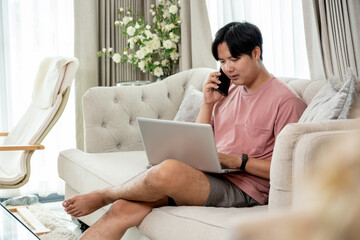 An Asian man in casual clothes using his laptop on a couch in the living room, working remotely from home or scrolling on website.