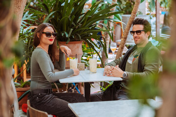 Young attractive couple wear sunglasses, drinking a cocktail while having a date sitting at the table in terrace bar with plants. Alcohol, beverage, drink concept.