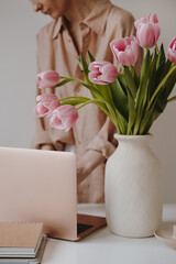 Lady boss workspace. Laptop computer, notebooks, pink tulip flowers in ceramic vase on a table....