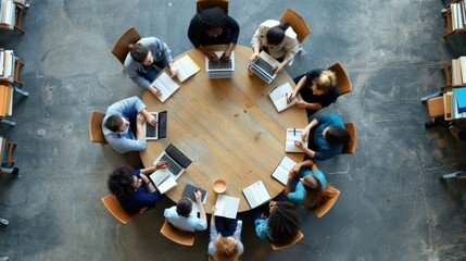 A diverse group of individuals engaged in a brainstorming session around a round wooden table, each using laptops and notepads for collaboration in a well-lit office space