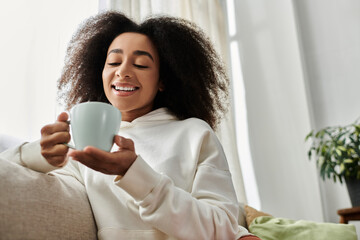 A cheerful young woman in a cozy outfit sips her drink while relaxing at home in her living room.