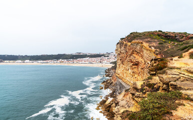 a stunning coastal view from atop a rugged cliff, overlooking a long stretch of sandy beach that reaches towards a quaint seaside town, under a muted sky.