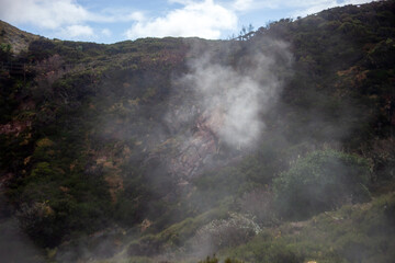 Sulfurous steam rises from the rocky landscape of Furnas de Enxofre, Terceira Island, Azores, Portugal. A unique geothermal site surrounded by lush green vegetation and rugged terrain.