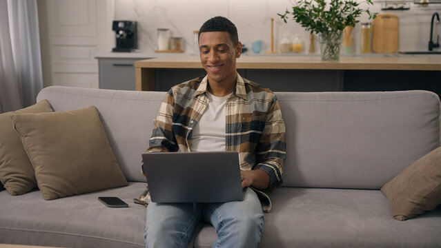 Happy smiling African American ethnic man working at home distant online work studying at sofa guy laptop using computer at couch male using pc playing internet video game use app shopping chatting