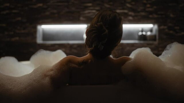 A relaxed woman enjoys a luxurious bubble bath in a dimly lit, modern bathroom setting.