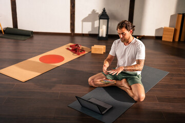 A young yoga instructor is conducting an online class while sitting on a yoga mat in his studio. He...