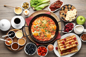 Tasty shakshouka in frying pan and other meals served for breakfast on wooden table, flat lay