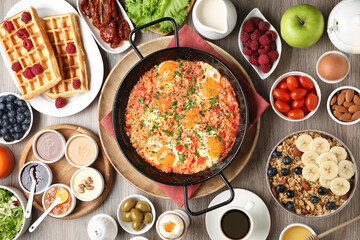 Tasty shakshouka in frying pan and other meals served for breakfast on wooden table, flat lay