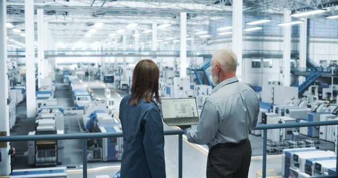 Two Multiethnic Engineers Standing on a Platform, Using Laptop Computer at an Electronics Factory. Machines are Undergoing Maintenance, Specialists Monitoring the Progress Through Online Software