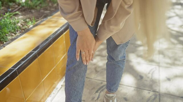 Young woman in casual attire holding her knee in apparent pain while standing outdoors on a sunny day.