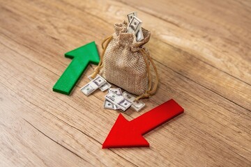 A burlap sack filled with a dollar bills between a green upward arrow and a red downward arrow....