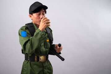 Asian male security officer in a green uniform and black cap, holding a baton, sipping a drink from a cup, taking a break during his duty. the plain white background