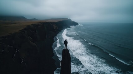 A solitary figure stands on a cliff overlooking a dramatic coastline, with waves crashing against the rocks under a cloudy sky.