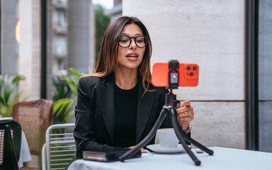 businesswoman records a video on her smartphone set up on a tripod, speaking professionally in an outdoor setting, blending technology and work