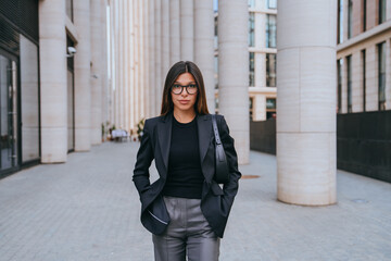 businesswoman in a black blazer and glasses stands confidently between modern city pillars, hands...