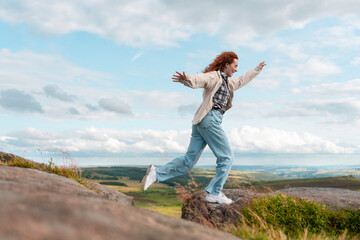 Fototapeta premium Joyful Woman Dancing on Rocky Outcrop Against a Backdrop of Blue Skies and Rolling Hills