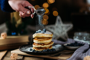 female hand sprinkles pancakes with powdered sugar, blueberry pancakes on a dark ceramic plate on a wooden background, breakfast, festive food