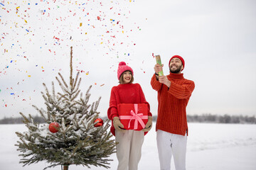 Man and woman have fun together, shooting confetti near Christmas tree outdoors. Young couple...