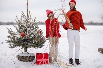Man and woman have fun together, shooting confetti near Christmas tree outdoors. Young couple...