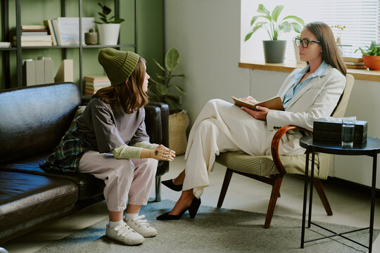 Therapist in white suit sitting opposite patient on couch during session discussing problems while patient gestures with hand in modern well-lit office