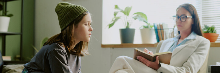 Young girl wearing beanie engaging in conversation with therapist holding notebook, seated in comfortable room with plants and sunlight filling space from window