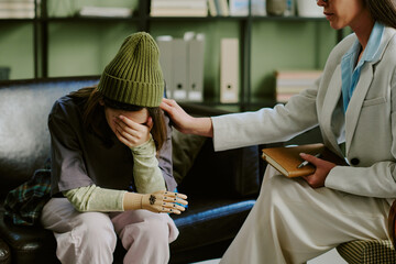 Woman with prosthetic arm sitting on couch with another woman offering support by patting her back...