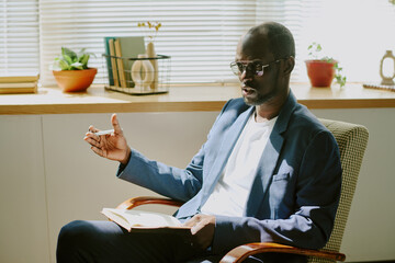 African American man sitting in chair near window holding book with bookshelf and potted plants in...