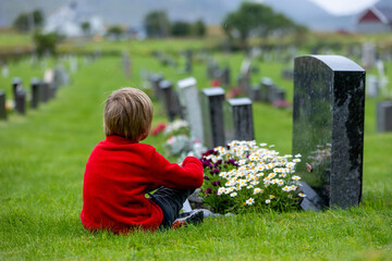 Sad little child, blond boy, standing in the rain on cemetery, sad person, mourning, rainy day