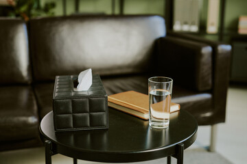 Tissue box, notebook, and glass of water placed on a round table next to a leather sofa, suggesting a cozy and organized living space