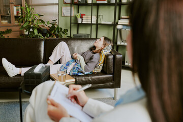 Client laying on couch in cozy office with therapist taking notes on clipboard and bookshelf in background creating inviting atmosphere