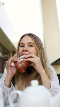 A beautiful blonde girl is eating a fresh powdered croissant at a caf&eacute; on summer terrace and smiling at the camera. girl is taking an appetizing bite of  croissant. Breakfast in Paris. Vertical video