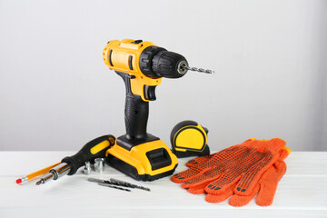 Cordless electric drill, gloves, bits, screwdriver and measuring tape on white wooden table