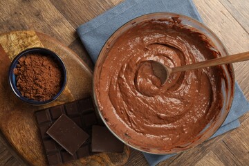 Chocolate dough in bowl, spoon and ingredients on wooden table, flat lay