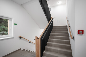 A modern interior staircase with wooden handrails, tiled steps, and an emergency exit sign. The space features clean white walls, a window, and no people are present.