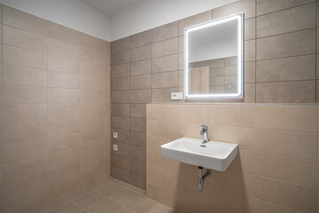 Minimalist bathroom with beige tiles, an illuminated mirror, and a sleek white sink. Clean design and contemporary look with subtle lighting.