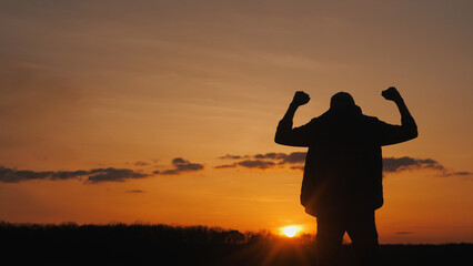 Fototapeta premium Silhouette of a young man in a victory gesture. Standing in a field at sunset