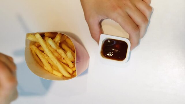 Woman's hands take French fries from a red paper package. She dips a piece into the bbq sauce and eats it. Top view of fast food and a white table in a cafe.