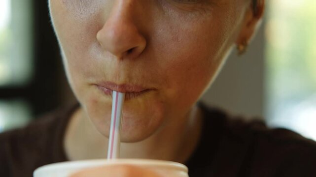 A young Caucasian woman with short haircut drinking soda from a paper cup using a straw in a cafe. Close-up of thoughtful girl drinking cola while sitting at cafe and smiling.