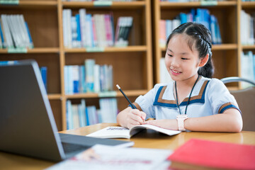 Young girl in a school uniform studying alone at a desk, writing in a notebook while referencing a laptop, smiling and focused, surrounded by bookshelves, capturing a moment of independent learning