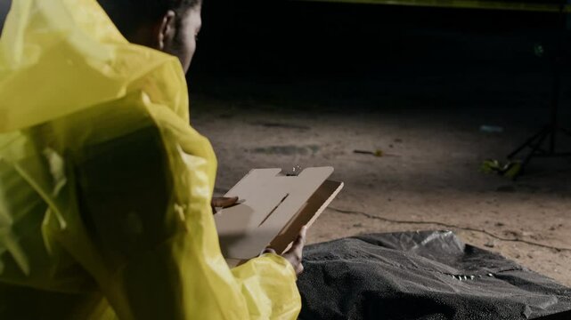 African American male detective in yellow raincoat squatting by murdered victim under plastic bag with criminals profile folder in hands while working on case on rainy night