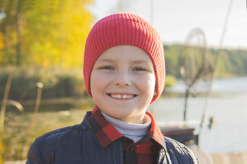 Portrait of a happy smiling boy in a red hat and jacket standing by an autumn lake with fishing gear and rods in the background.