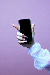 Hand of spooky, white ghost holding smartphone with blank screen against pastel violet background. Concept of season sale, promotion, celebration, web. Negative space to insert text. Ad